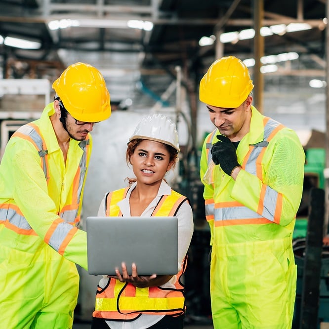 Three Gen Z factory employees wearing safety gear reviewing workplace information together on a laptop.
