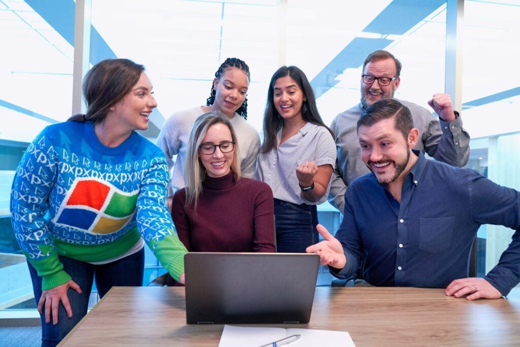 Six coworkers gathered around a laptop in an office celebrating together.