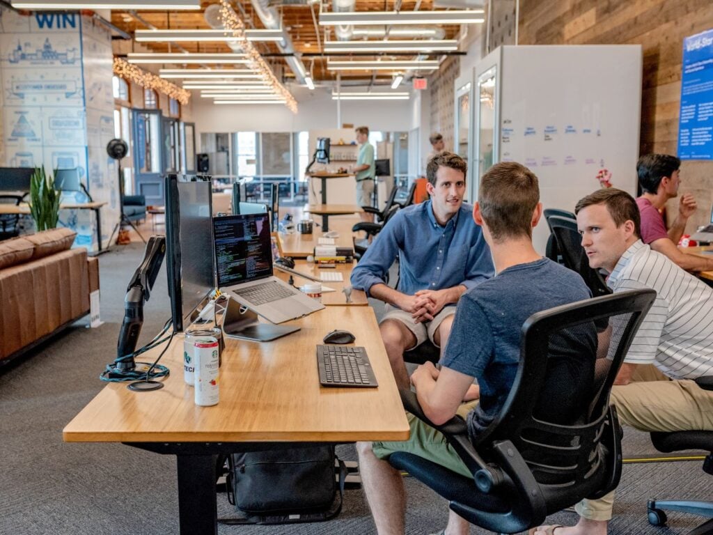 Employees in a modern office having a discussion while seated at desks with laptops.